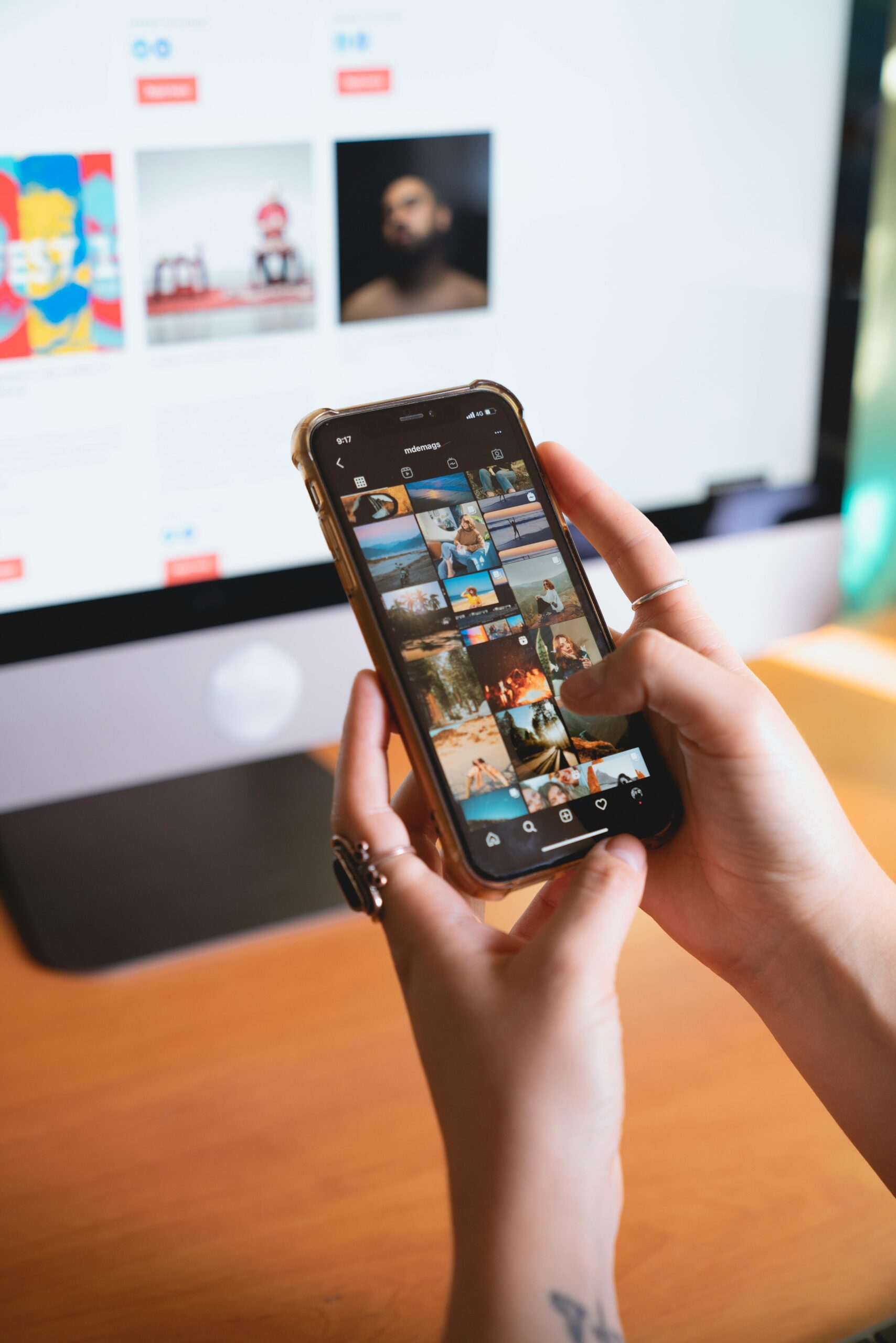 Close-up of a person browsing a smartphone with an Instagram feed, with a computer monitor in the background.
